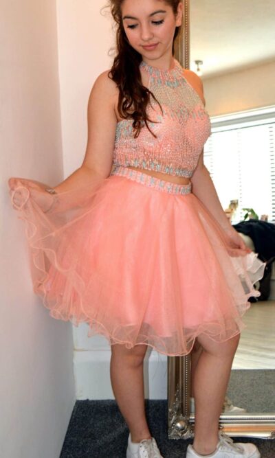 Teenage model wearing a Blush Prom Dress Style 89 in coral. Two piece that features a completely encrusted jewel neckline and top, matching beaded waist, and chiffon skirt from Silhouette London, Girls party dress specialists in London.